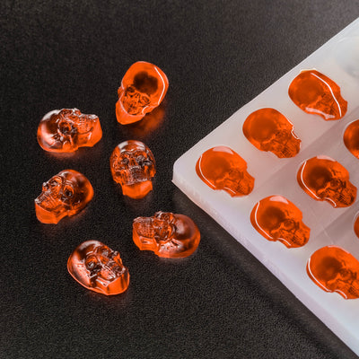 Skull-shaped orange gummy candies on a black surface with a silicone mold.