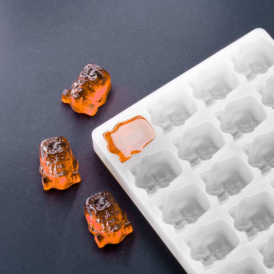 A white silicone candy mold with the shape of a Frankenstein head, with three orange candies made from the mold beside it.